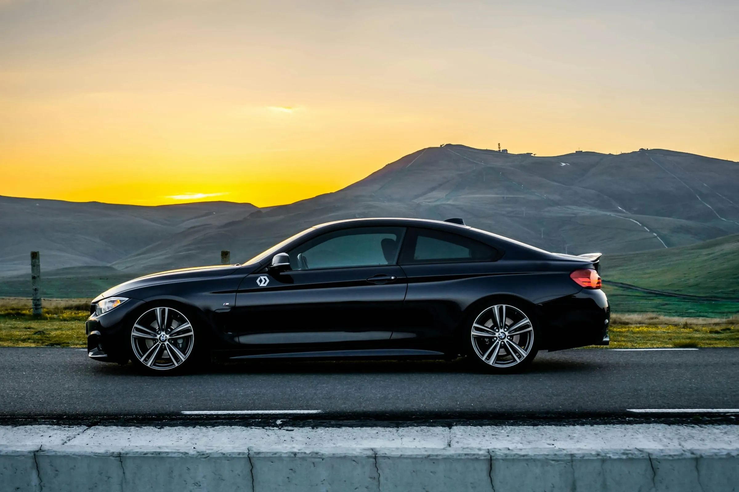 A black BMW coupe parked on an empty road at sunset, rolling hills behind it, the paint reflecting the sky