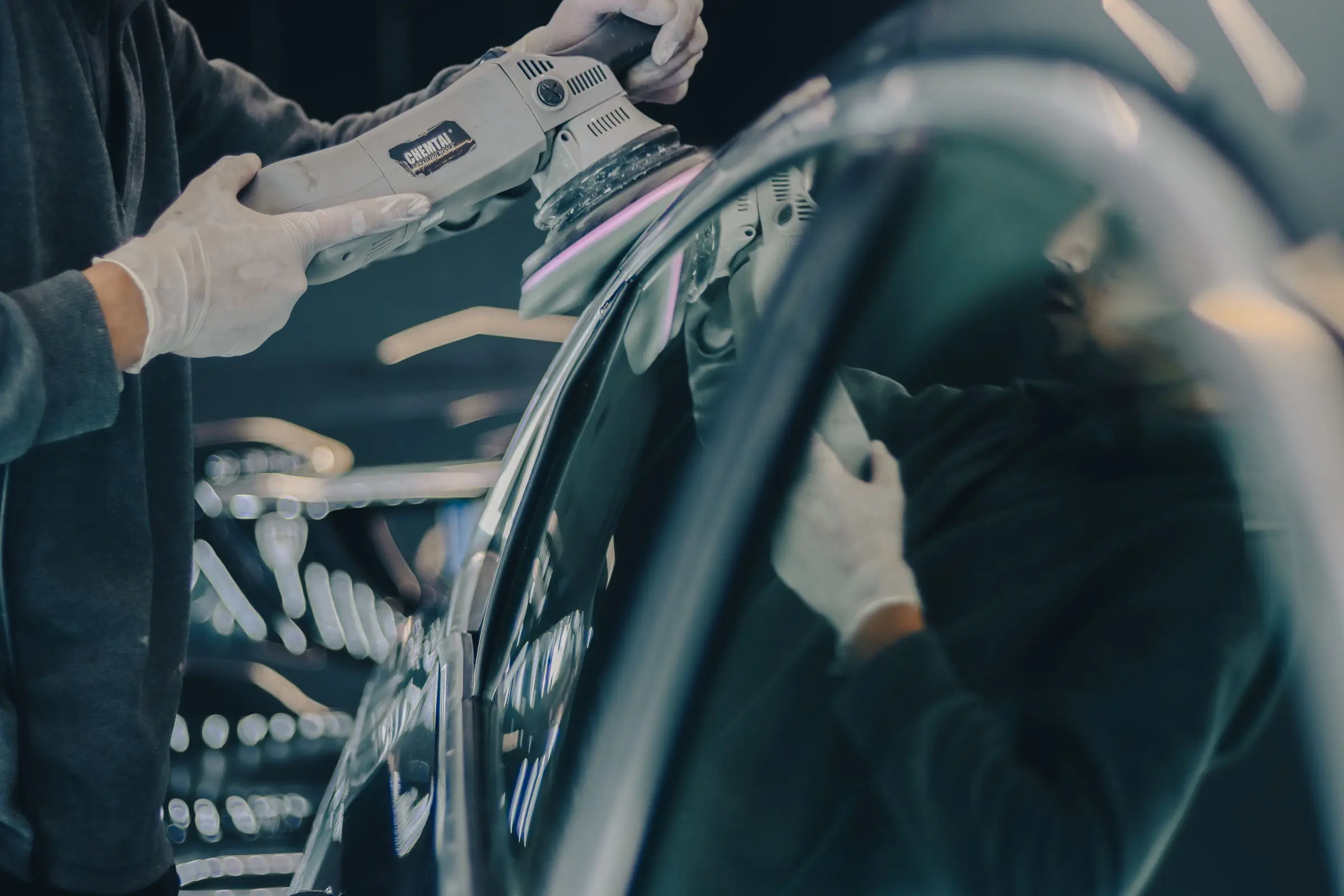 A gloved detailer working a dual-action polisher along the top of a dark car door in a workshop.