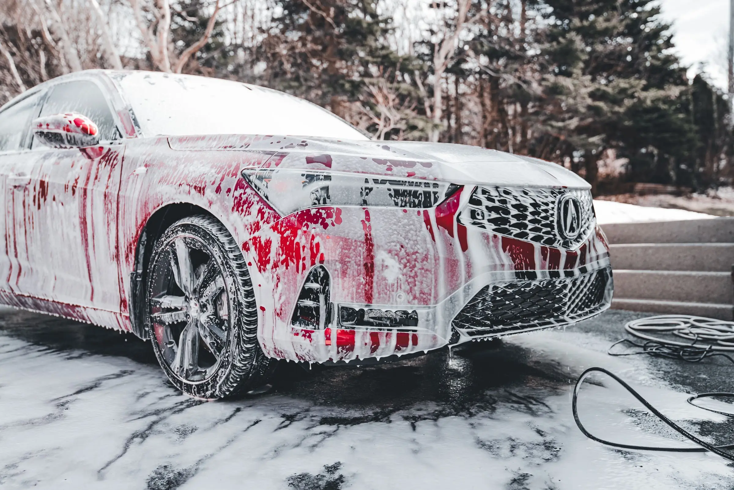 A red car covered in thick white soapy foam during an outdoor wash.